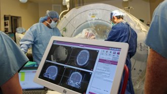 A computer monitor shows various brain imagery, while two surgeons operating on a person are visible in the background.