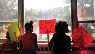 Children holding "Clean our water," "Don't be mean, keep our water clean" and other protst signs in front of a window.